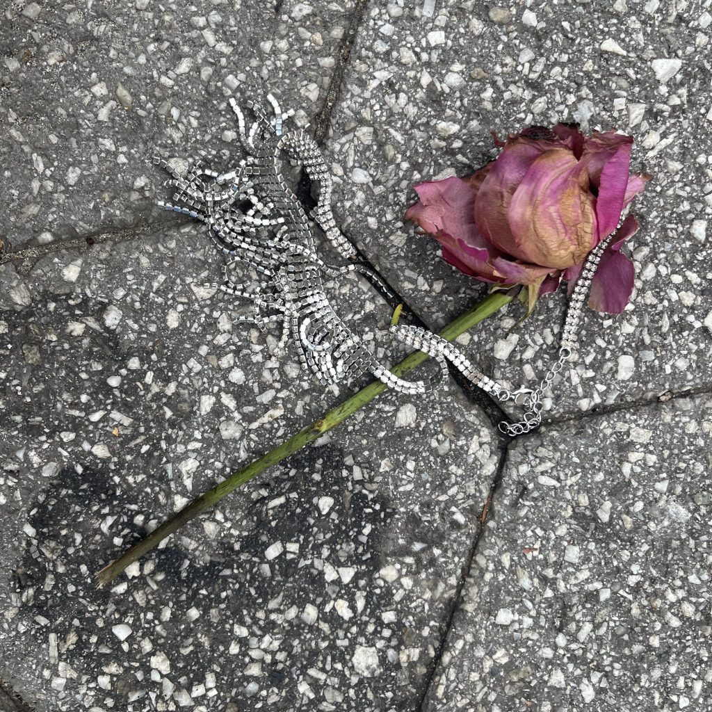 A dead rose wrapped in a sparkly necklace sits on concrete.